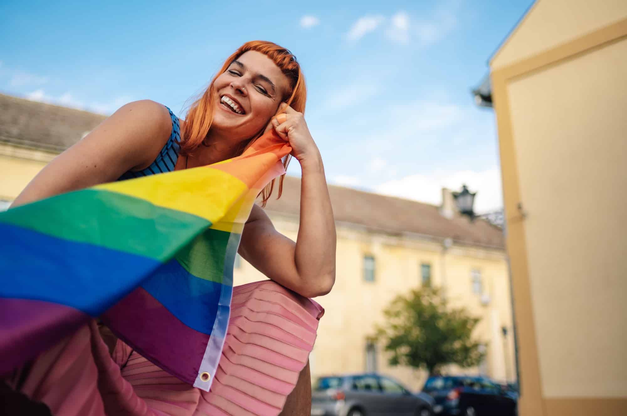 Young redhead woman with lgbt rainbow flag smiling in urban environment